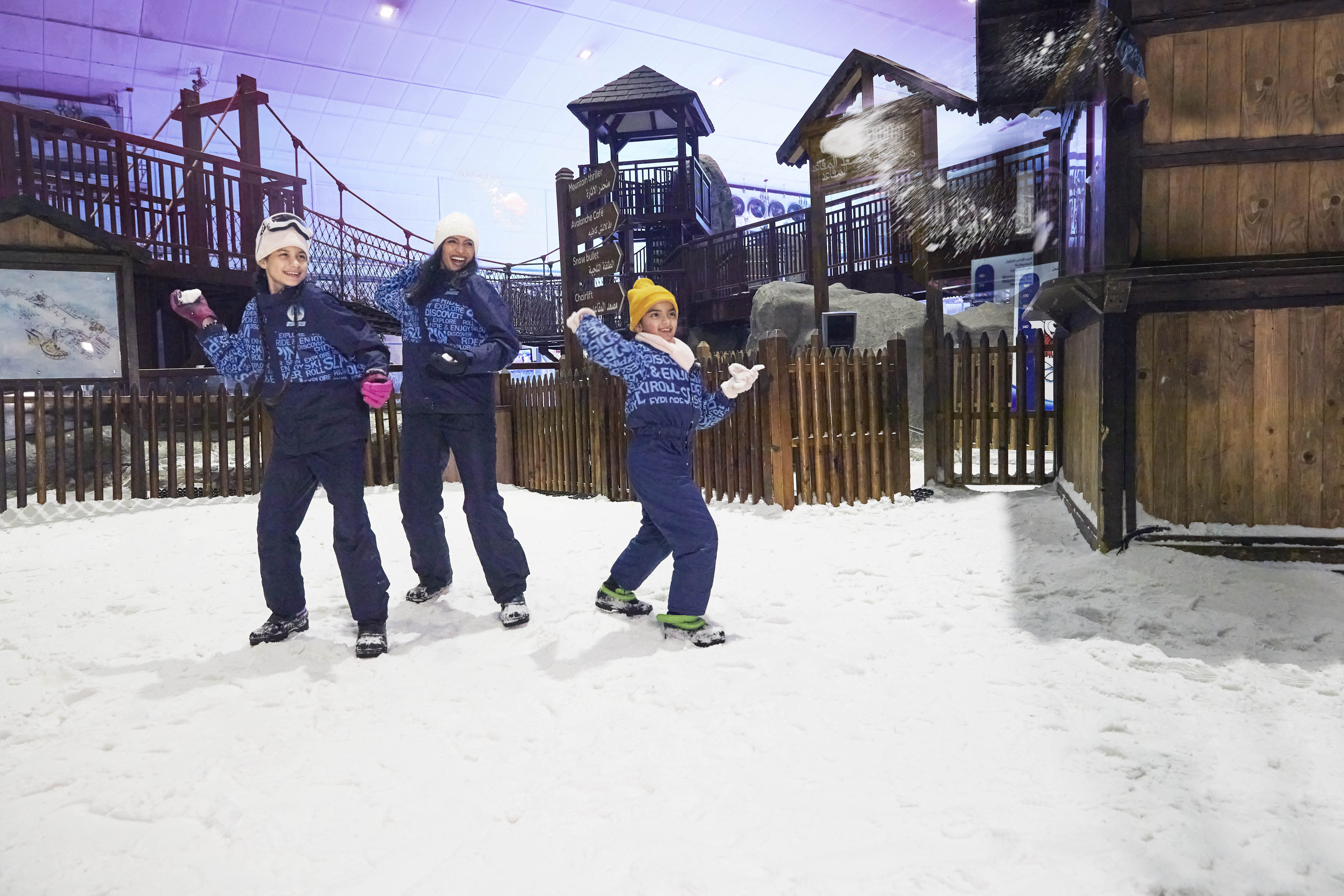 Families enjoy a snowy indoor play area, throwing snowballs and laughing near wooden structures in a winter wonderland.