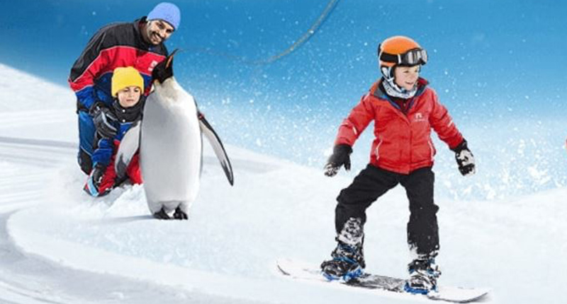 Family enjoying a snowy adventure, with a child snowboarding and another riding with a penguin, set against a vibrant blue...