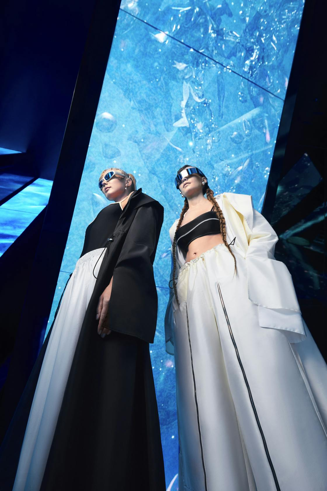 Two models showcase futuristic fashion in striking black and white outfits, set against a vibrant blue digital backdrop.