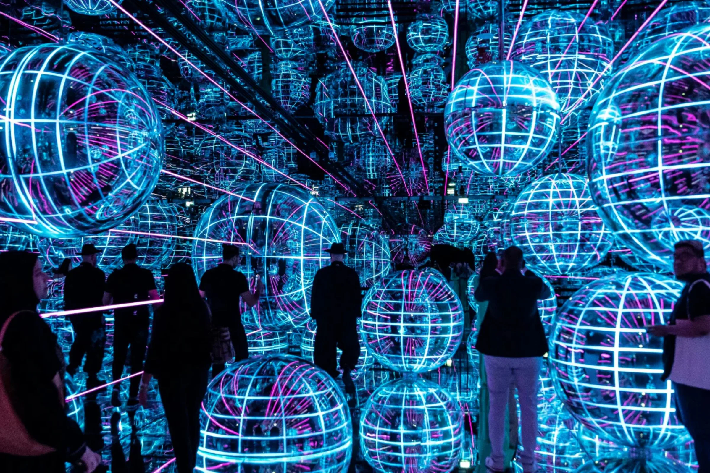 Visitors explore a mesmerizing installation of glowing blue and pink spherical lights in a mirrored space.