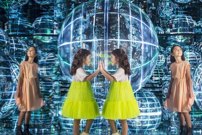 Two young girls in colorful dresses interact with a glowing, mirrored orb in a vibrant, illuminated art installation.