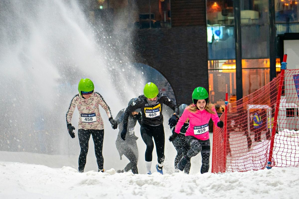 Participants in colorful helmets race through artificial snow during an indoor skiing event, creating a fun winter atmosph...