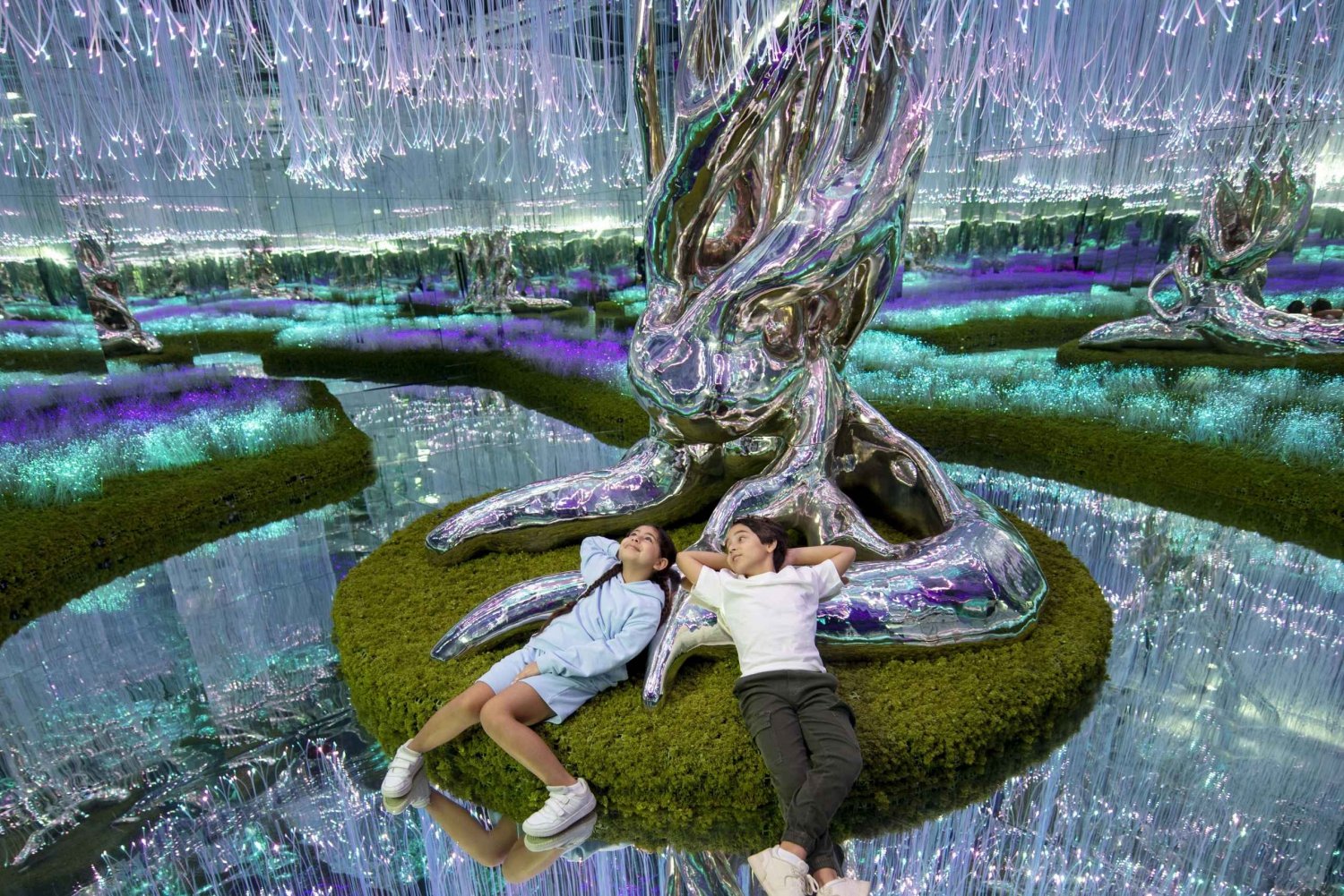Two children relax on a vibrant green moss patch, surrounded by shimmering mirrors and colorful lights in a stunning art i...